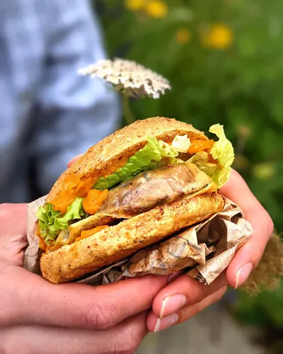 Un burger SUPERWASTE tenu à la main, devant un massif de fleurs.