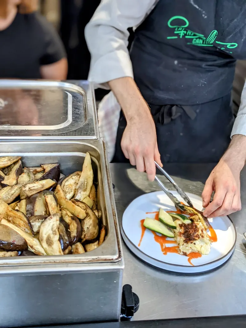 En cuisine à La Fabuleuse Cantine : une cheffe dresse une assiette à côté d’un bac de légumes rôtis.